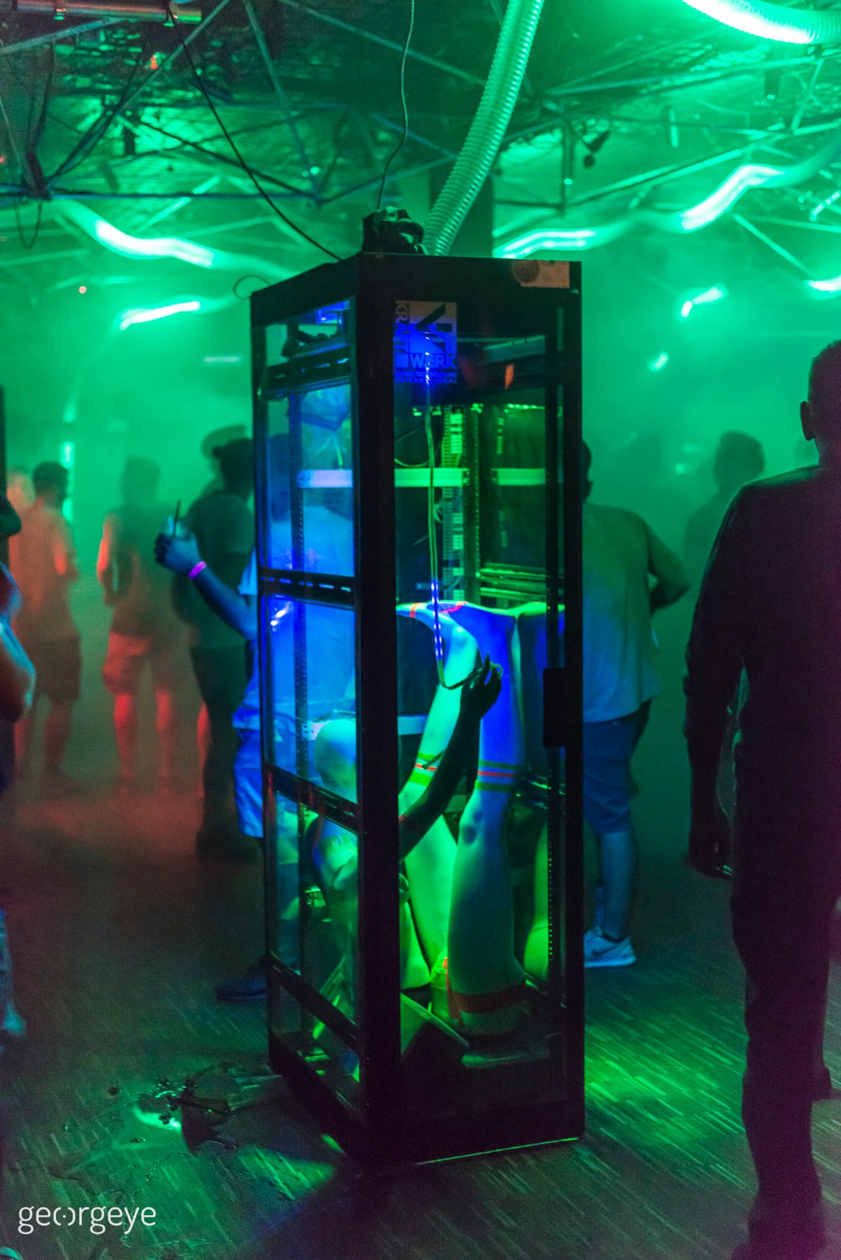 A mildly filled dark club with green lights on the ceiling and in the middle a server rack with LED strips and a broken mannequin on its floor